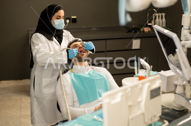الفحص الطبي و تقديم الخدمات الطبية للمرضى , مفهوم الرعاية والعناية بالمرضى ،  صورة مقربة لطبيبة عربية خليجيه سعودية ترتدي القفازات والكمامة الطبية تقوم بفحص أسنان المريض ، مفهوم الطب والرعاية الصحية ، إجراء الفحوصات الدورية للعناية بالصحة الفموية ، شاب عربي خليجي سعودي يرتدي الزي التقليدي يجلس في العيادة السنية وينظر الى الكاميرا , شاب عماني يجلس على كرسي العلاج  .