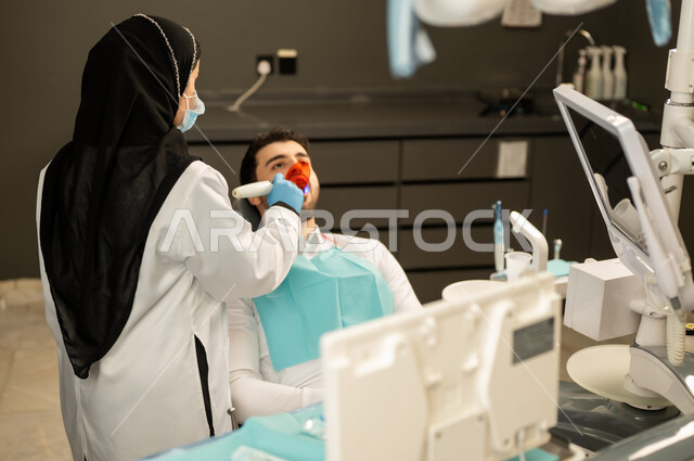 مفهوم الرعاية والعناية بالمرضى ، الفحص الطبي و تقديم الخدمات الطبية للمرضى ,  صورة مقربة لطبيبة عربية خليجيه سعودية ترتدي القفازات والكمامة الطبية تقوم بفحص أسنان المريض ، مفهوم الطب والرعاية الصحية ، إجراء الفحوصات الدورية للعناية بالصحة الفموية ، شاب عربي خليجي سعودي يرتدي الزي التقليدي يجلس في العيادة السنية وينظر الى الكاميرا , شاب عماني يجلس على كرسي العلاج  .