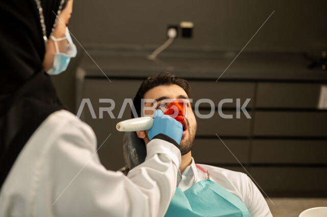 المعدات اللازمة لإجراء الفحوصات الطبية ، معاينة الحالات المرضية و تشخيصها ، صورة مقربة لطبيبة عربية خليجيه سعودية ترتدي القفازات والكمامة الطبية تقوم بفحص أسنان المريض ، مفهوم الطب والرعاية الصحية ، إجراء الفحوصات الدورية للعناية بالصحة الفموية ، شاب عربي خليجي سعودي يرتدي الزي التقليدي يجلس في العيادة السنية وينظر الى الكاميرا , شاب عماني يجلس على كرسي العلاج  .