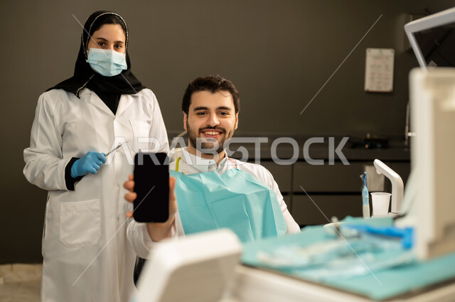 دمج التقنيات الحديثة والتكنولوجيا في مجال الطب ,  صورة مقربة لطبيبة عربية خليجيه سعودية ترتدي القفازات والكمامة الطبية تقوم بفحص أسنان المريض ، مفهوم الطب والرعاية الصحية ، إجراء الفحوصات الدورية للعناية بالصحة الفموية ، شاب عربي خليجي سعودي يرتدي الزي التقليدي يجلس في العيادة السنية ويعرض شاشة سوداء فارغة للجوال , شاب عماني يجلس على كرسي العلاج  .