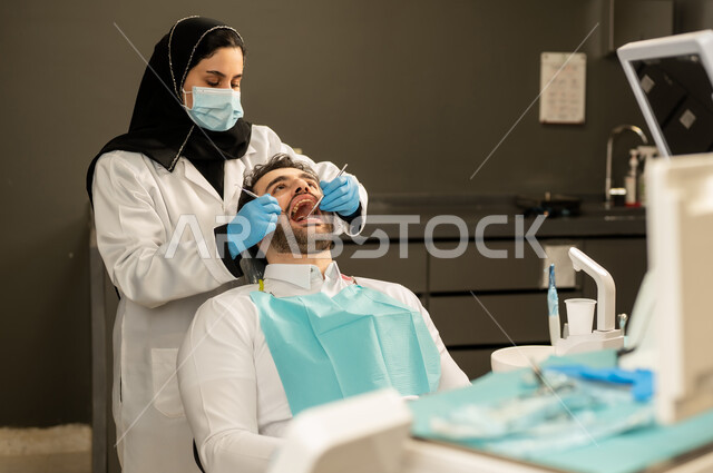 استخدام الادوات اللازمة لإجراء الفحوصات الطبيه ,معالجة التسوس و النخر, شاب عربي خليجي سعودي يرتدي الزي التقليدي يجلس في العيادة السنية ويفتح فمه لتشخيص المرض, صورة مقربة لطبيبة عربية خليجيه سعودية ترتدي القفازات والكمامة الطبية تقوم بفحص أسنان المريض ، شاب عماني على كرسي العلاج  .