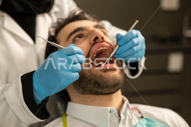 Oral and dental surgery, providing health services in specialized centers, conducting medical examinations, a young Saudi Arabian Gulf man wearing traditional clothing sitting in a dental clinic and opening his mouth to diagnose a disease, a close-up of a female Saudi Arabian Gulf doctor wearing gloves and a medical mask examining a patient's teeth, a young Omani man sitting on a treatment chair.