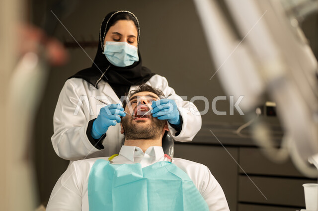معالجة التسوس و النخر, استخدام الادوات اللازمة لإجراء الفحوصات الطبيه ,شاب عربي خليجي سعودي يرتدي الزي التقليدي يجلس في العيادة السنية ويفتح فمه لتشخيص المرض, صورة مقربة لطبيبة عربية خليجيه سعودية ترتدي القفازات والكمامة الطبية تقوم بفحص أسنان المريض ، شاب عماني على كرسي العلاج  .