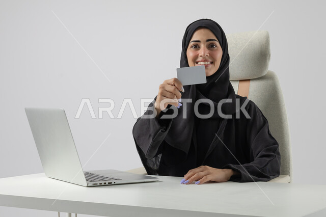 عرض موك اب ابيض صغير، وظيفة نسائية مكتبية سعودية، ادارة وانجاز المهام باستخدام جهاز تقني حديث ومتطور, بورتريه مقرب إمرأة عربية خليجية سعودية مبتسمة ترتدي الحجاب والعباءة تعرض بطاقة تعريفية، العمل على الحاسوب المحمول, ايماءات وتعابير السرور والسعادة، خلفية بيضاء