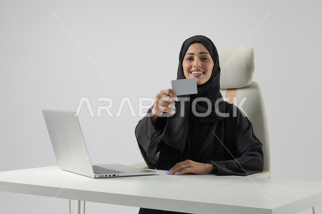 عرض موك اب ابيض صغير، وظيفة نسائية مكتبية سعودية، ادارة وانجاز المهام باستخدام جهاز تقني حديث ومتطور, بورتريه مقرب إمرأة عربية خليجية سعودية مبتسمة ترتدي الحجاب والعباءة تعرض بطاقة تعريفية، العمل على الحاسوب المحمول, ايماءات وتعابير السرور والسعادة، خلفية بيضاء
