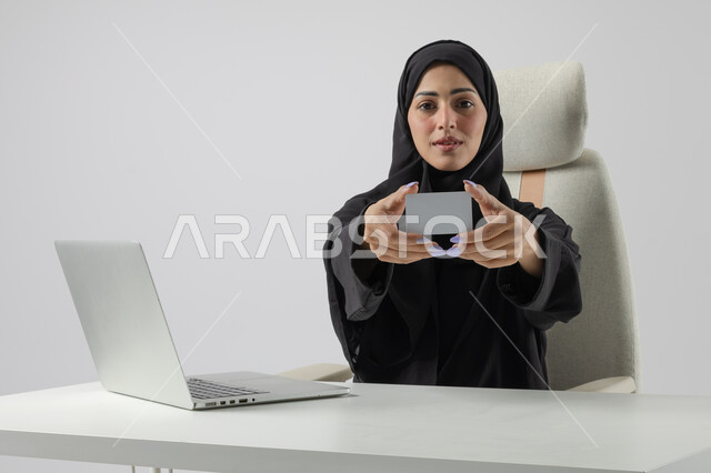 عرض موك اب ابيض صغير، وظيفة نسائية مكتبية سعودية، ادارة وانجاز المهام باستخدام جهاز تقني حديث ومتطور, بورتريه مقرب إمرأة عربية خليجية سعودية مبتسمة ترتدي الحجاب والعباءة تعرض بطاقة تعريفية، العمل على الحاسوب المحمول, ايماءات وتعابير السرور والسعادة، خلفية بيضاء