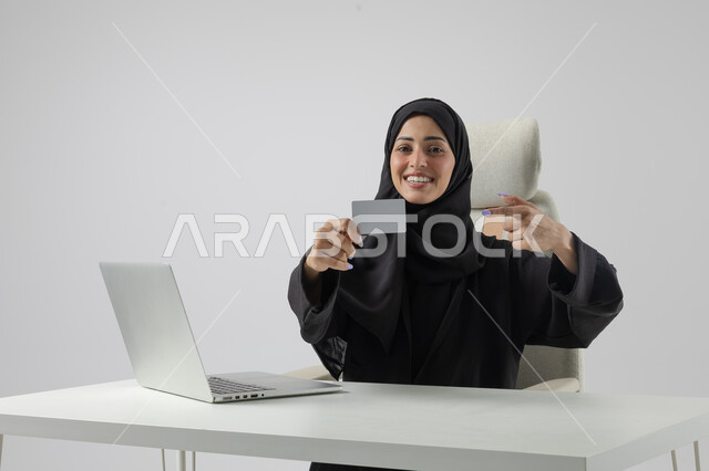 عرض موك اب ابيض صغير، وظيفة نسائية مكتبية سعودية، ادارة وانجاز المهام باستخدام جهاز تقني حديث ومتطور, بورتريه مقرب إمرأة عربية خليجية سعودية مبتسمة ترتدي الحجاب والعباءة تعرض بطاقة تعريفية، العمل على الحاسوب المحمول, ايماءات وتعابير السرور والسعادة، خلفية بيضاء