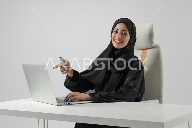 Pointing with index finger, Saudi female office job, managing and completing tasks using modern and advanced technical device, close-up portrait of a smiling Saudi Arabian Gulf woman wearing hijab and abaya pointing at laptop, gestures and expressions of pleasure and happiness, white background