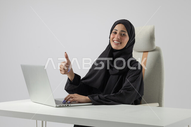 تعابير القبول والرضا والاعجاب، وظيفة نسائية مكتبية سعودية، ادارة وانجاز المهام باستخدام جهاز تقني حديث ومتطور, بورتريه مقرب إمرأة عربية خليجية سعودية مبتسمة ترتدي الحجاب والعباءة تعمل على الحاسوب المحمول, ايماءات السرور والسعادة، خلفية بيضاء