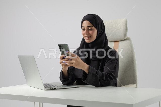 استخدام الهاتف المحمول، وظيفة نسائية مكتبية سعودية، ادارة وانجاز المهام باستخدام جهاز تقني حديث ومتطور, بورتريه مقرب إمرأة عربية خليجية سعودية مبتسمة ترتدي الحجاب والعباءة تعمل على الحاسوب المحمول, ايماءات وتعابير السرور والسعادة، خلفية بيضاء