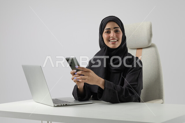 استخدام الهاتف المحمول، وظيفة نسائية مكتبية سعودية، ادارة وانجاز المهام باستخدام جهاز تقني حديث ومتطور, بورتريه مقرب إمرأة عربية خليجية سعودية مبتسمة ترتدي الحجاب والعباءة تعمل على الحاسوب المحمول, ايماءات وتعابير السرور والسعادة، خلفية بيضاء