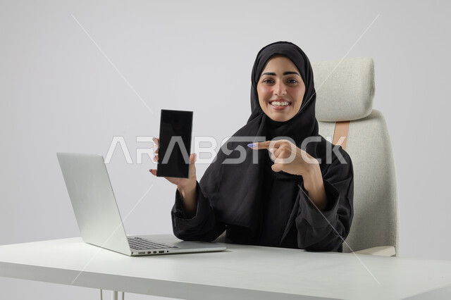 عرض شاشة سوداء فارغة على الهاتف المحمول، وظيفة نسائية مكتبية سعودية، ادارة وانجاز المهام باستخدام جهاز تقني حديث ومتطور, بورتريه مقرب إمرأة عربية خليجية سعودية مبتسمة ترتدي الحجاب والعباءة تعمل على الحاسوب المحمول, ايماءات وتعابير السرور والسعادة، خلفية بيضاء