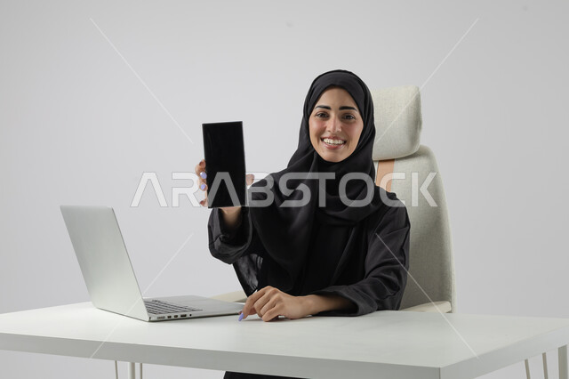 عرض شاشة سوداء فارغة على الهاتف المحمول، وظيفة نسائية مكتبية سعودية، ادارة وانجاز المهام باستخدام جهاز تقني حديث ومتطور, بورتريه مقرب إمرأة عربية خليجية سعودية مبتسمة ترتدي الحجاب والعباءة تعمل على الحاسوب المحمول, ايماءات وتعابير السرور والسعادة، خلفية بيضاء