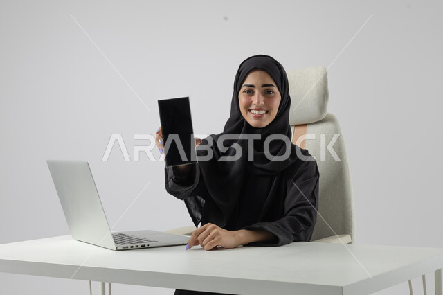 عرض شاشة سوداء فارغة على الهاتف المحمول، وظيفة نسائية مكتبية سعودية، ادارة وانجاز المهام باستخدام جهاز تقني حديث ومتطور, بورتريه مقرب إمرأة عربية خليجية سعودية مبتسمة ترتدي الحجاب والعباءة تعمل على الحاسوب المحمول, ايماءات وتعابير السرور والسعادة، خلفية بيضاء
