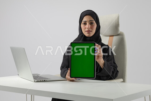 عرض شاشة خضراء كروما فارغة على جهاز التابلت، وظيفة نسائية مكتبية سعودية، ادارة وانجاز المهام باستخدام جهاز تقني حديث ومتطور, بورتريه مقرب إمرأة عربية خليجية سعودية مبتسمة ترتدي الحجاب والعباءة تعمل على الحاسوب المحمول, تعابير السرور والسعادة، خلفية بيضاء 