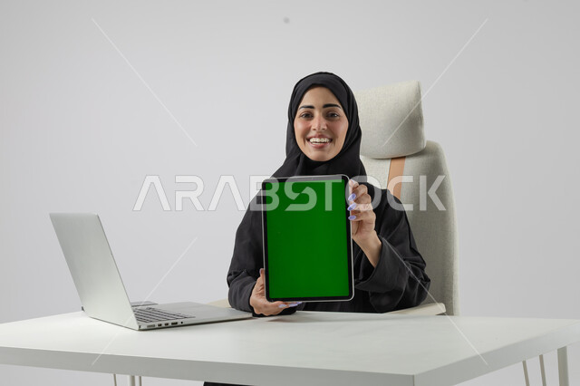  عرض شاشة خضراء كروما فارغة على جهاز التابلت، وظيفة نسائية مكتبية سعودية، ادارة وانجاز المهام باستخدام جهاز تقني حديث ومتطور, بورتريه مقرب إمرأة عربية خليجية سعودية مبتسمة ترتدي الحجاب والعباءة تعمل على الحاسوب المحمول, تعابير السرور والسعادة، خلفية بيضاء 