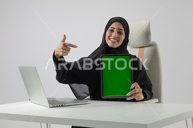  عرض شاشة خضراء كروما فارغة على جهاز التابلت، وظيفة نسائية مكتبية سعودية، ادارة وانجاز المهام باستخدام جهاز تقني حديث ومتطور, بورتريه مقرب إمرأة عربية خليجية سعودية مبتسمة ترتدي الحجاب والعباءة تعمل على الحاسوب المحمول, تعابير السرور والسعادة، خلفية بيضاء 