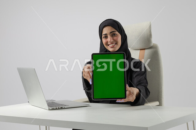 عرض شاشة خضراء كروما فارغة على جهاز التابلت، وظيفة نسائية مكتبية سعودية، ادارة وانجاز المهام باستخدام جهاز تقني حديث ومتطور, بورتريه مقرب إمرأة عربية خليجية سعودية مبتسمة ترتدي الحجاب والعباءة تعمل على الحاسوب المحمول, تعابير السرور والسعادة، خلفية بيضاء 