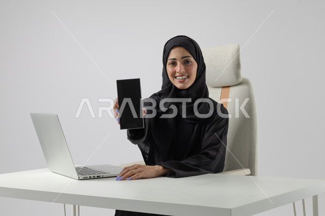 عرض شاشة سوداء فارغة على الهاتف المحمول، وظيفة نسائية مكتبية سعودية، ادارة وانجاز المهام باستخدام جهاز تقني حديث ومتطور, بورتريه مقرب إمرأة عربية خليجية سعودية مبتسمة ترتدي الحجاب والعباءة تعمل على الحاسوب المحمول, ايماءات وتعابير السرور والسعادة، خلفية بيضاء
