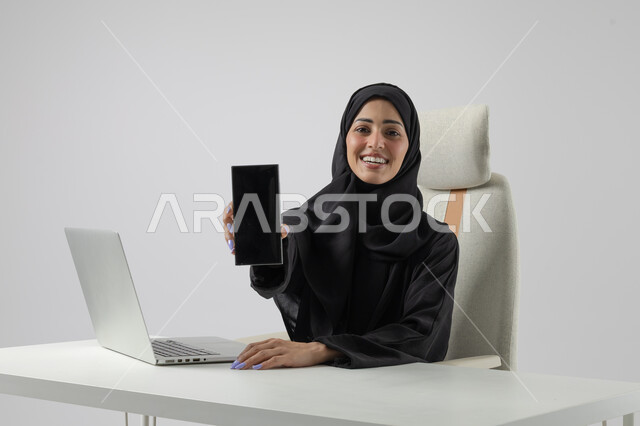 عرض شاشة سوداء فارغة على الهاتف المحمول، وظيفة نسائية مكتبية سعودية، ادارة وانجاز المهام باستخدام جهاز تقني حديث ومتطور, بورتريه مقرب إمرأة عربية خليجية سعودية مبتسمة ترتدي الحجاب والعباءة تعمل على الحاسوب المحمول, ايماءات وتعابير السرور والسعادة، خلفية بيضاء