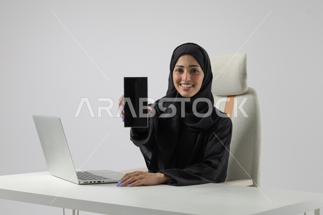 عرض شاشة سوداء فارغة على الهاتف المحمول، وظيفة نسائية مكتبية سعودية، ادارة وانجاز المهام باستخدام جهاز تقني حديث ومتطور, بورتريه مقرب إمرأة عربية خليجية سعودية مبتسمة ترتدي الحجاب والعباءة تعمل على الحاسوب المحمول, ايماءات وتعابير السرور والسعادة، خلفية بيضاء