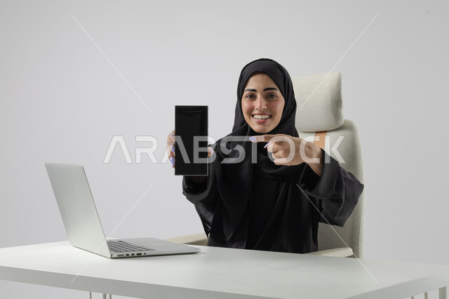 عرض شاشة سوداء فارغة على الهاتف المحمول، وظيفة نسائية مكتبية سعودية، ادارة وانجاز المهام باستخدام جهاز تقني حديث ومتطور, بورتريه مقرب إمرأة عربية خليجية سعودية مبتسمة ترتدي الحجاب والعباءة تعمل على الحاسوب المحمول, ايماءات وتعابير السرور والسعادة، خلفية بيضاء