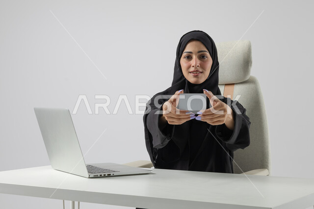 عرض موك اب ابيض صغير، وظيفة نسائية مكتبية سعودية، ادارة وانجاز المهام باستخدام جهاز تقني حديث ومتطور, بورتريه مقرب إمرأة عربية خليجية سعودية مبتسمة ترتدي الحجاب والعباءة تعرض بطاقة تعريفية، العمل على الحاسوب المحمول, ايماءات وتعابير السرور والسعادة، خلفية بيضاء
