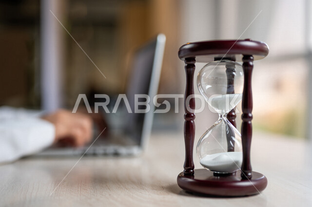صورة مقربة ليد رجل عربي خليجي سعودي يستخدم الحاسوب المحمول، تحديد موعد العمل من خلال الساعة رملية، انجاز المهام بدقة وكفاءة، ادارة الجدول الزمني، مفهوم ادارة الوقت