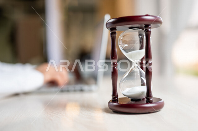 صورة مقربة ليد رجل عربي خليجي سعودي يستخدم الحاسوب المحمول، تحديد موعد العمل من خلال الساعة رملية، انجاز المهام بدقة وكفاءة، ادارة الجدول الزمني، مفهوم ادارة الوقت