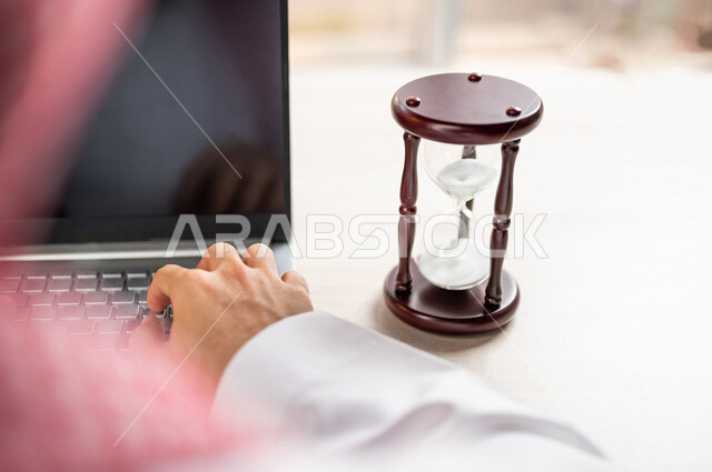 صورة مقربة ليد رجل عربي خليجي سعودي يستخدم الحاسوب المحمول، تحديد موعد العمل من خلال الساعة رملية، انجاز المهام بدقة وكفاءة، ادارة الجدول الزمني، مفهوم ادارة الوقت