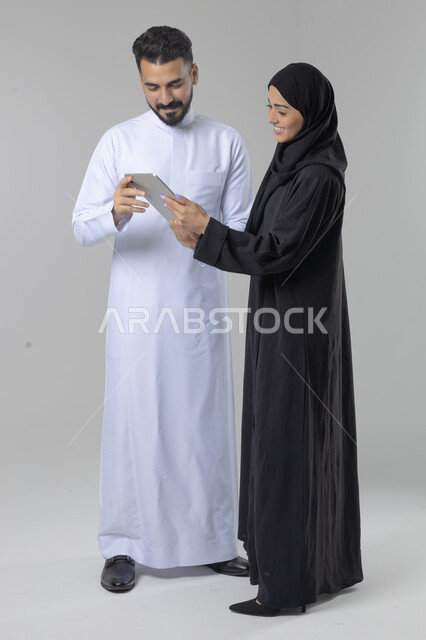 تبادل الأفكار والخبرات لتحقيق أهداف الشركة، العمل الجماعي وروح الفريق الواحد، بورتريه لزميلين عربيين سعوديين خليجيين يقومان بمناقشة و تنظيم الاعمال بإستخدام جهاز التابليت، دمج العمل مع التقنية والتكنولوجيا، صورة الجسم بالطول الكامل، خلفية بيضاء