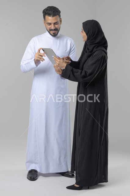 تبادل الأفكار والخبرات لتحقيق أهداف الشركة، العمل الجماعي وروح الفريق الواحد، بورتريه لزميلين عربيين سعوديين خليجيين يقومان بمناقشة و تنظيم الاعمال بإستخدام جهاز التابليت، دمج العمل مع التقنية والتكنولوجيا، صورة الجسم بالطول الكامل، خلفية بيضاء