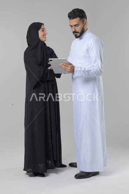 تبادل الأفكار والخبرات لتحقيق أهداف الشركة، العمل الجماعي وروح الفريق الواحد، بورتريه لزميلين عربيين سعوديين خليجيين يقومان بمناقشة و تنظيم الاعمال بإستخدام جهاز التابليت، دمج العمل مع التقنية والتكنولوجيا، صورة الجسم بالطول الكامل، خلفية بيضاء