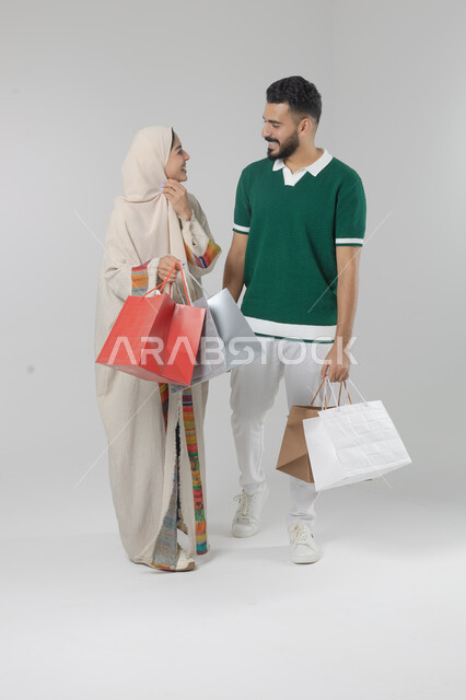 Taking advantage of seasonal offers and discounts, expressions and gestures of pleasure and happiness, buying gifts and supplies, portrait of a Saudi Arabian Gulf couple holding shopping bags in their hands, full body, white background