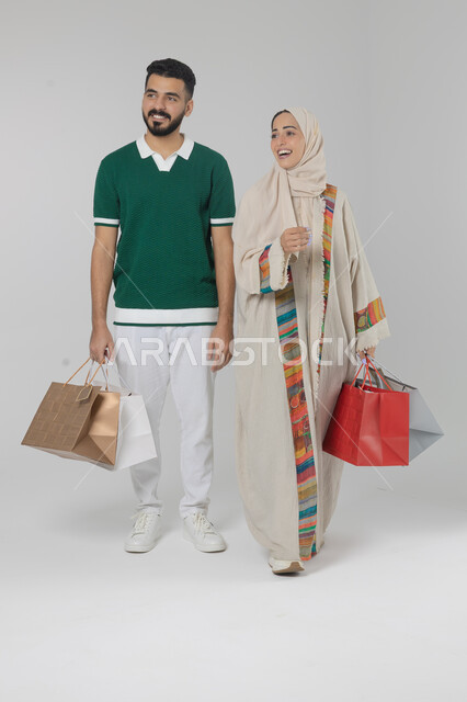 Taking advantage of seasonal offers and discounts, expressions and gestures of pleasure and happiness, buying gifts and supplies, portrait of a Saudi Arabian Gulf couple holding shopping bags in their hands, full body, white background