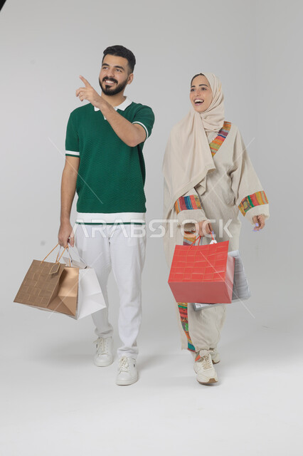 الاستفادة من العروض والتخفيضات الموسميه، تعابير وايماءات السرور والسعادة، شراء الهدايا والمستلزمات، بورتريه لزوجين عربيين خليجيين سعوديين يحملان بيديهما أكياس التسوق، الجسم بالطول الكامل، خلفية بيضاء