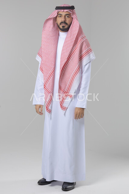 الاعتناء والاهتمام بالمظهر الخارجي، مفهوم الاناقة والرجولة، النظر الى شيء ما، بورتريه لشاب عربي خليجي سعودي يرتدي الثوب التقليدي والشماغ يقف باستقامة بتعابير السرور والثقة بالنفس، الجسم بالطول الكامل، خلفية بيضاء