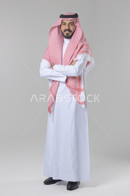 الاعتناء والاهتمام بالمظهر الخارجي، مفهوم الاناقة والرجولة، التقاط صور شخصية للوثائق الرسمية، بورتريه لشاب عربي خليجي سعودي يرتدي الثوب التقليدي والشماغ يقف بأيدي متقاطعة ينظر الى الكاميرا بتعابير السرور والثقة بالنفس، الجسم بالطول الكامل، خلفية بيضاء