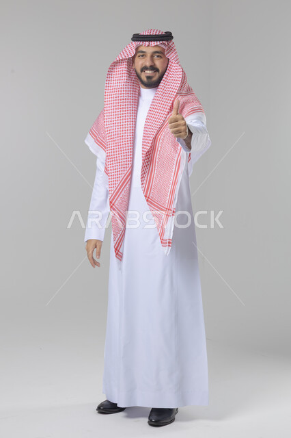  الإمتياز والتميز والنجاح، بورتريه لرجل عربي خليجي سعودي يرتدي الثوب التقليدي والشماغ ويرفع أصبع الإبهام لأعلى، إيماءات السعادة والإعجاب والموافقة، حركة أصبع تدل على القبول والرضا، الجسم بالطول الكامل، خلفية بيضاء 