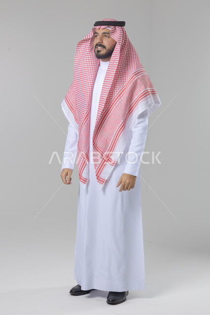 الاعتناء والاهتمام بالمظهر الخارجي، مفهوم الاناقة والرجولة، النظر الى شيء ما، بورتريه لشاب عربي خليجي سعودي يرتدي الثوب التقليدي والشماغ يقف باستقامة بتعابير السرور والثقة بالنفس، الجسم بالطول الكامل، خلفية بيضاء
