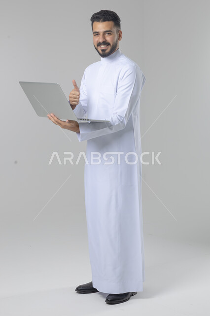 استخدام التقنية الحديثه المتطورة في الحياة العملية، تعابير وايماءات الاندماج والسرور، بورتريه لشاب عربي خليجي سعودي مبتسم يرتدي الثوب التقليدي يقوم بالعمل على الحاسب المحمول، صورة الجسم بالطول الكامل، خلفية بيضاء