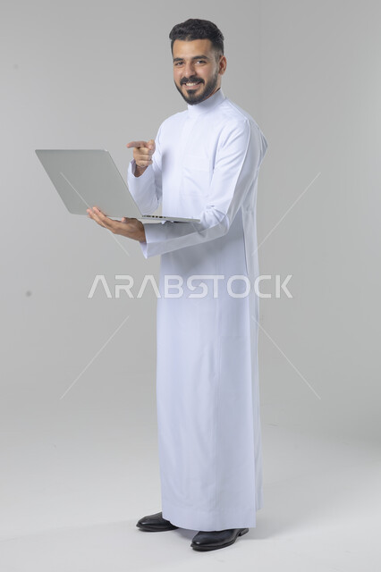 استخدام التقنية الحديثه المتطورة في الحياة العملية، تعابير وايماءات الاندماج والسرور، بورتريه لشاب عربي خليجي سعودي مبتسم يرتدي الثوب التقليدي يقوم بالعمل على الحاسب المحمول، صورة الجسم بالطول الكامل، خلفية بيضاء