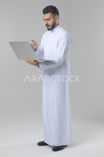 Using modern advanced technology in practical life, expressions and gestures of integration and pleasure, portrait of a smiling young Saudi Arabian Gulf man wearing traditional thobe working on a laptop, full body image, white background