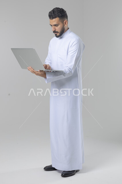 Using modern advanced technology in practical life, expressions and gestures of integration and pleasure, portrait of a smiling young Saudi Arabian Gulf man wearing traditional thobe working on a laptop, full body image, white background