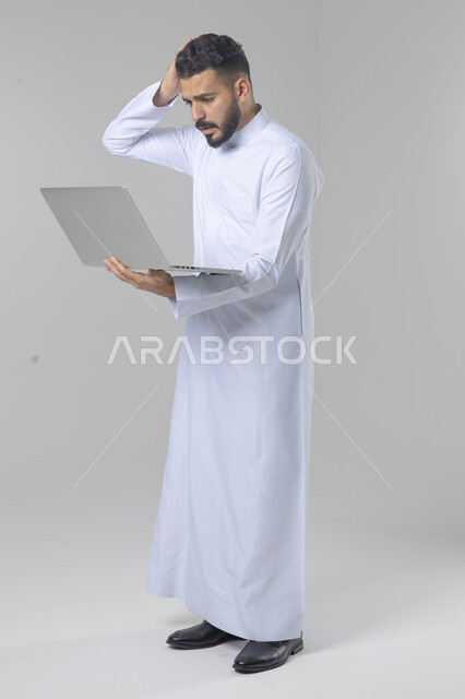استخدام التقنية الحديثه المتطورة في الحياة العملية، تعابير وايماءات الاندماج والسرور، بورتريه لشاب عربي خليجي سعودي مبتسم يرتدي الثوب التقليدي يقوم بالعمل على الحاسب المحمول، صورة الجسم بالطول الكامل، خلفية بيضاء