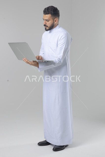 Using modern advanced technology in practical life, expressions and gestures of integration and pleasure, portrait of a smiling young Saudi Arabian Gulf man wearing traditional thobe working on a laptop, full body image, white background