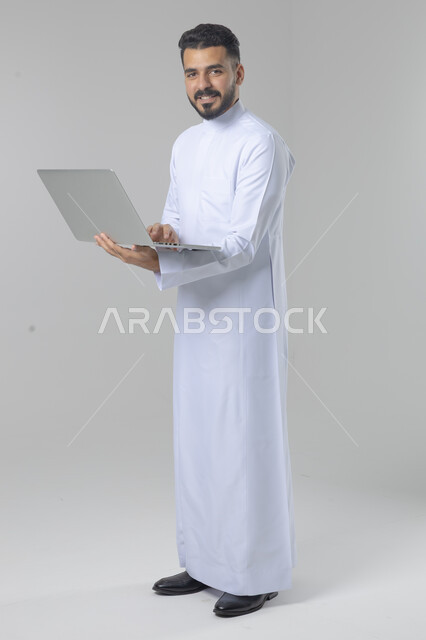 استخدام التقنية الحديثه المتطورة في الحياة العملية، تعابير وايماءات الاندماج والسرور، بورتريه لشاب عربي خليجي سعودي مبتسم يرتدي الثوب التقليدي يقوم بالعمل على الحاسب المحمول، صورة الجسم بالطول الكامل، خلفية بيضاء