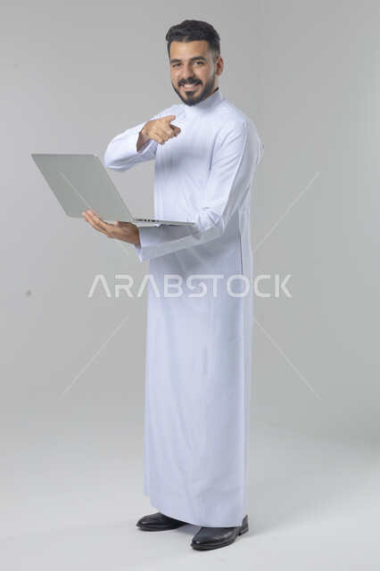 Using modern advanced technology in practical life, expressions and gestures of integration and pleasure, portrait of a smiling young Saudi Arabian Gulf man wearing traditional thobe working on a laptop, full body image, white background