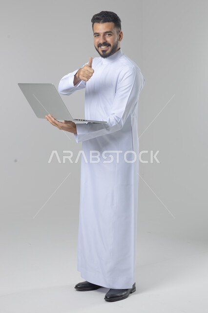 Using modern advanced technology in practical life, expressions and gestures of integration and pleasure, portrait of a smiling young Saudi Arabian Gulf man wearing traditional thobe working on a laptop, full body image, white background