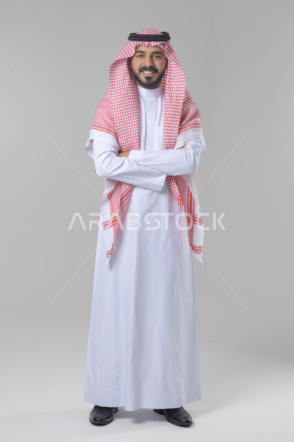 الاعتناء والاهتمام بالمظهر الخارجي، مفهوم الاناقة والرجولة، التقاط صور شخصية للوثائق الرسمية، بورتريه لشاب عربي خليجي سعودي يرتدي الثوب التقليدي والشماغ يقف بأيدي متقاطعة ينظر الى الكاميرا بتعابير السرور والثقة بالنفس، الجسم بالطول الكامل، خلفية بيضاء
